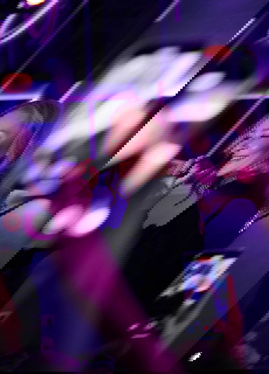 Исполнительница поёт в микрофон на сцене бара, в кадре размытые смартфоны публики и фиолетово-розовая концертная подсветка, атмосфера живого выступления.