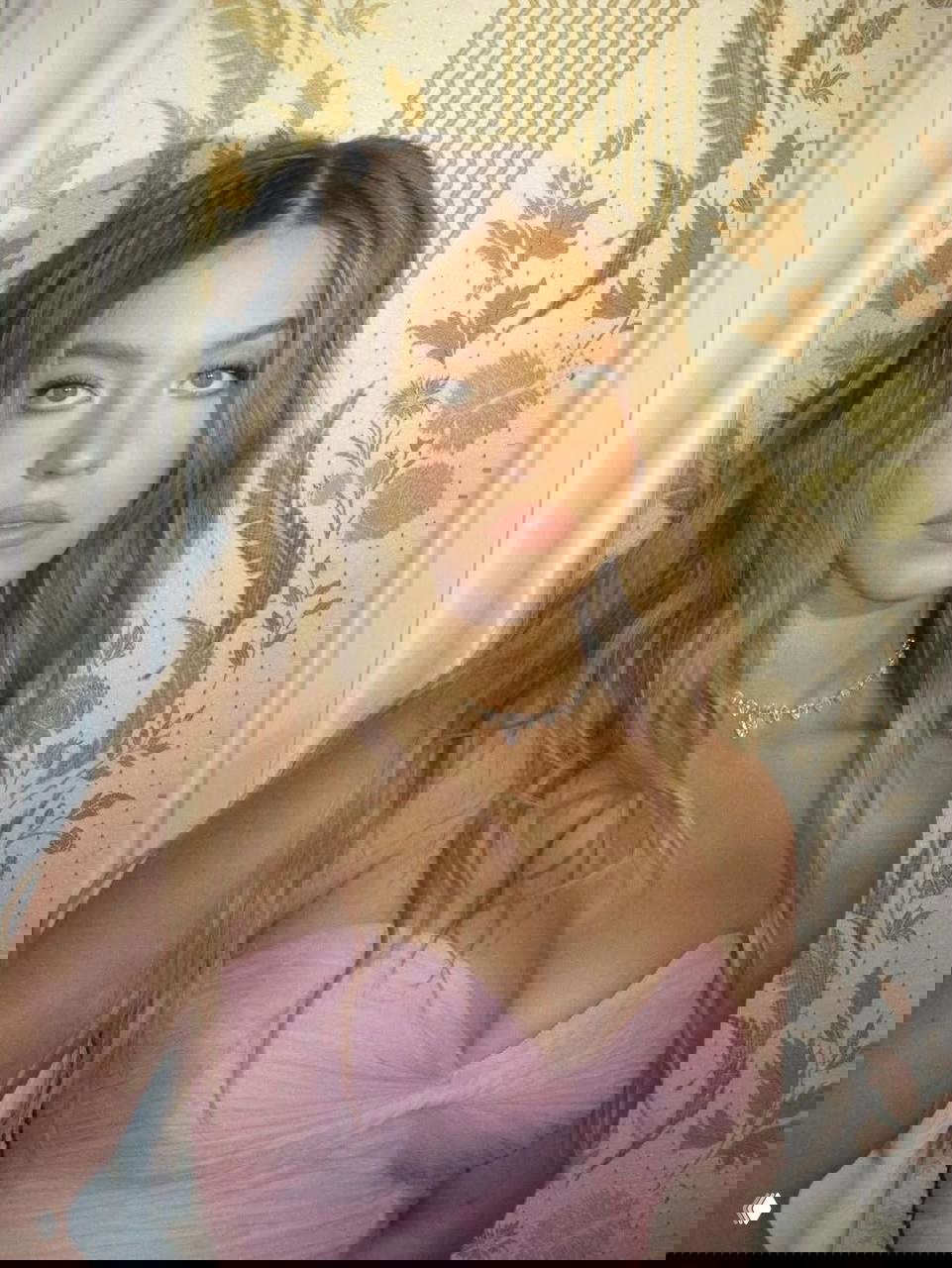 Портрет молодой женщины в розовом платье на фоне узорных обоев; интерьерная съёмка, крупный план и естественный макияж.