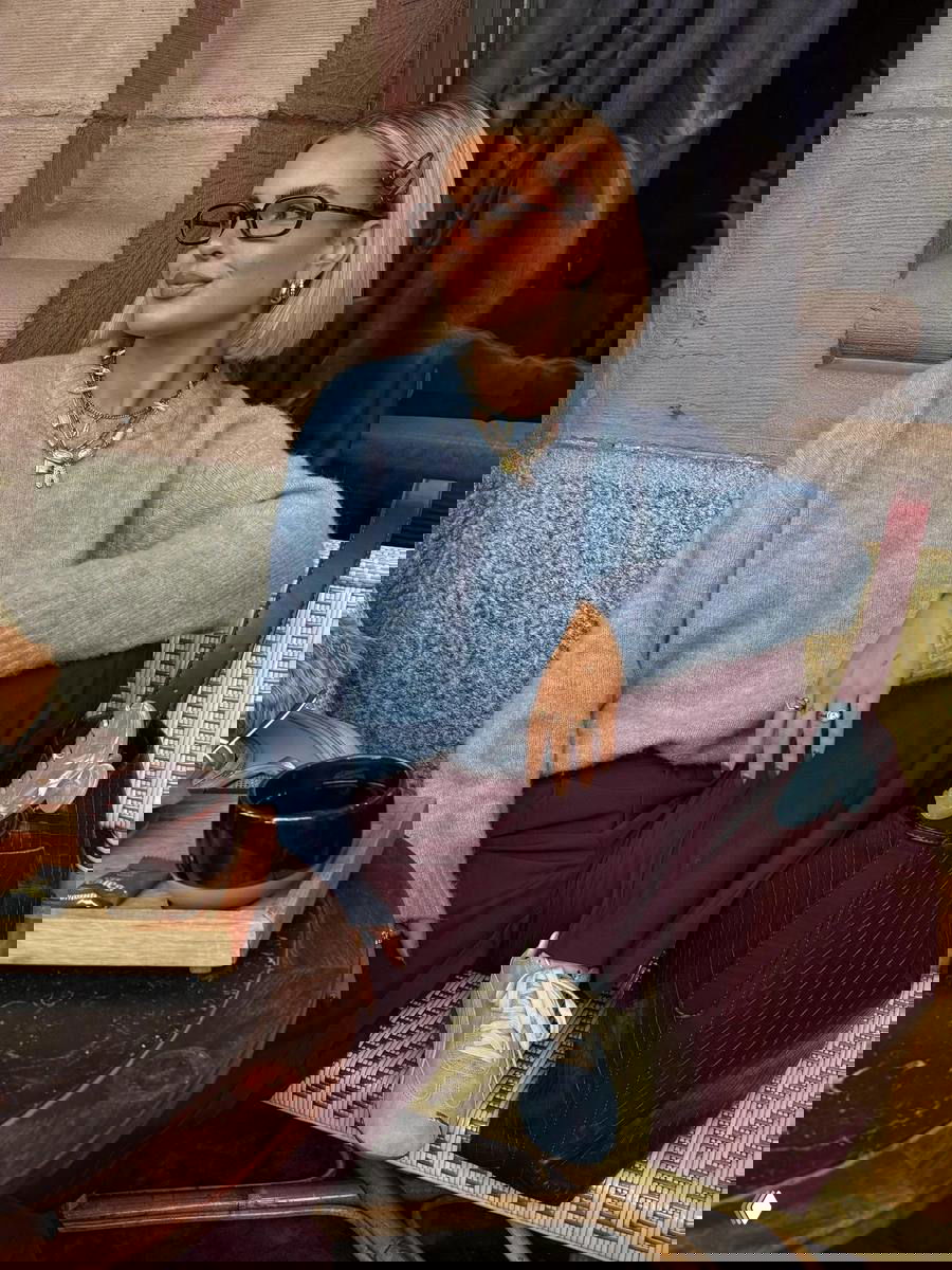 Сидящая девушка в нежно-голубом шерстяном свитере и бордовых брюках, ретро-кеды в светло-голубом и большая бордовая кожаная сумка.