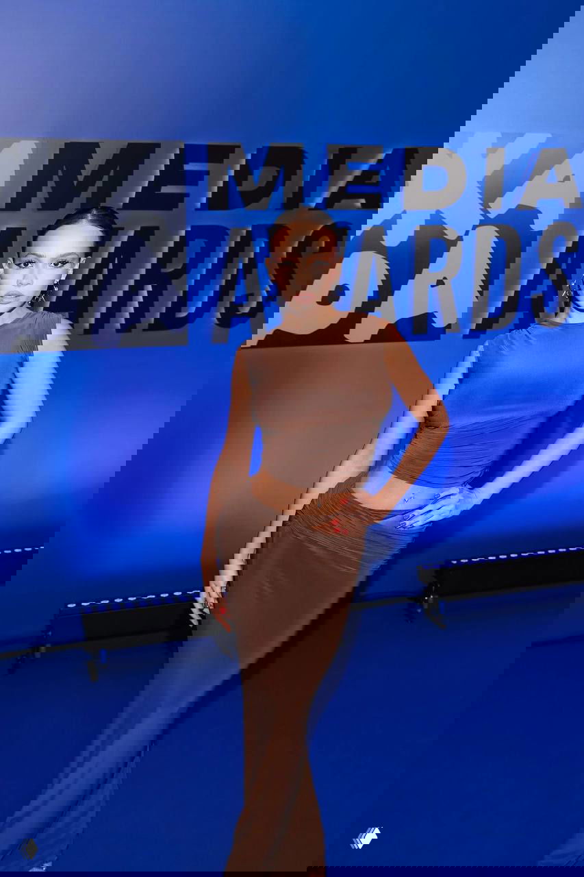 Молодая женщина в коричневом платье позирует на синем фоне у баннера «Media Awards» на профессиональном медиа-ивенте.