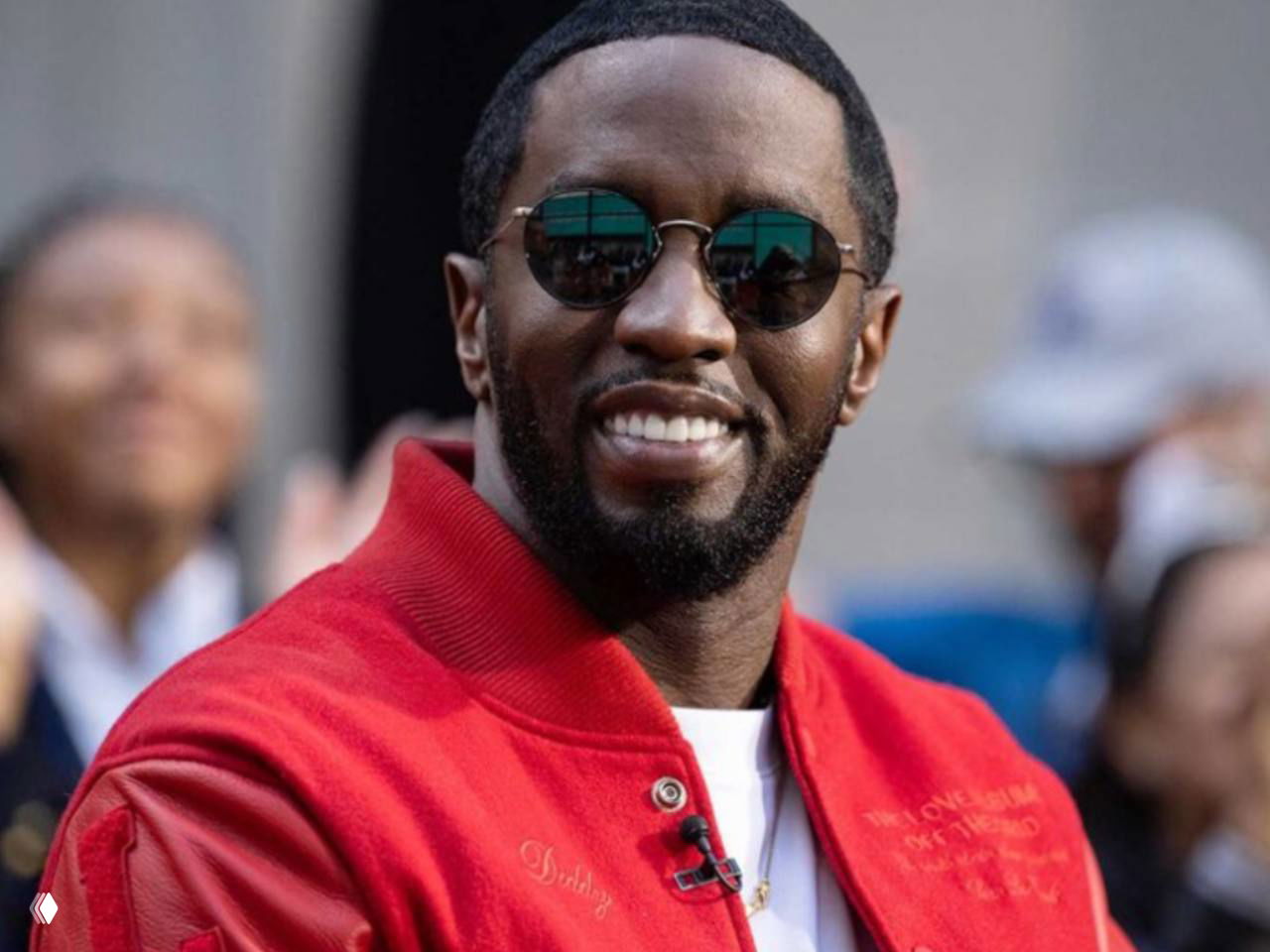 Человек в красной куртке и круглых солнечных очках на публичном мероприятии, крупный план лица, на заднем плане размытая толпа и охрана.