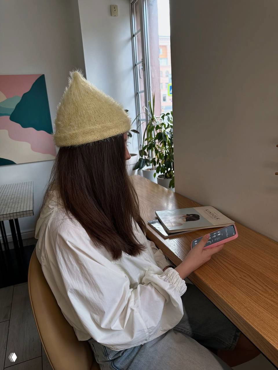 Женщина в светлой остроконечной вязаной шапке, сидит за деревянным столом у окна, держит телефон и журнал в уютном интерьерe.