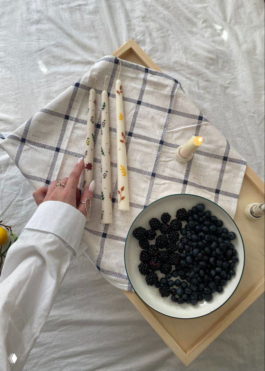 Снимок нескольких расписных свечей на клетчатой ткани рядом с чашей черники и ежевики; рука аккуратно раскладывает свечи в кадре