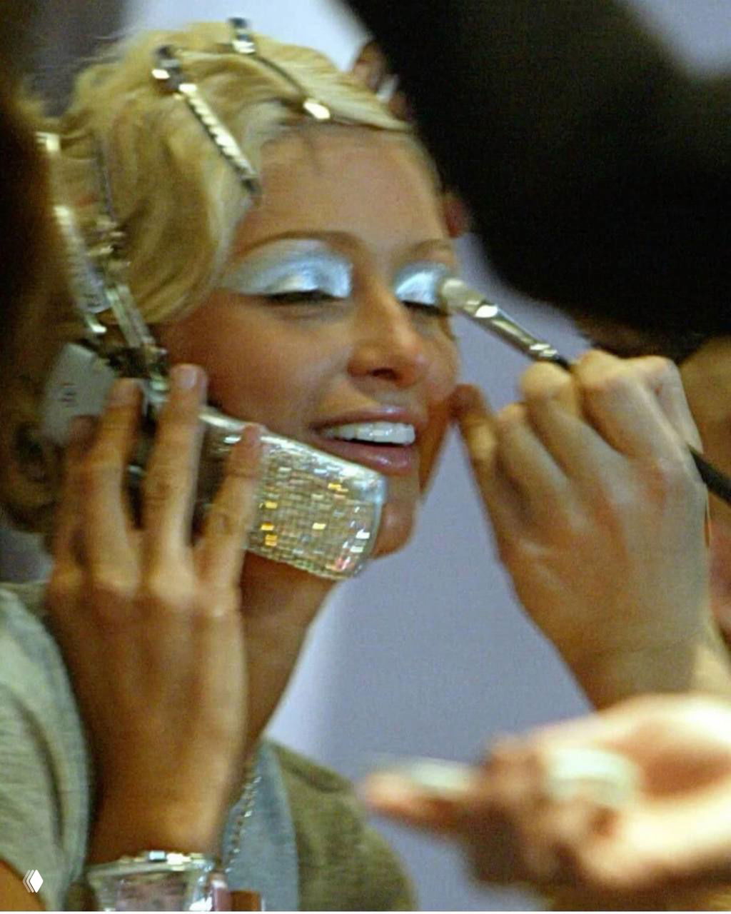 Крупный кадр: женщина во время нанесения серебристых теней и блеска, видны кисточки и аксессуары, акцент на металлике в макияже глаз.