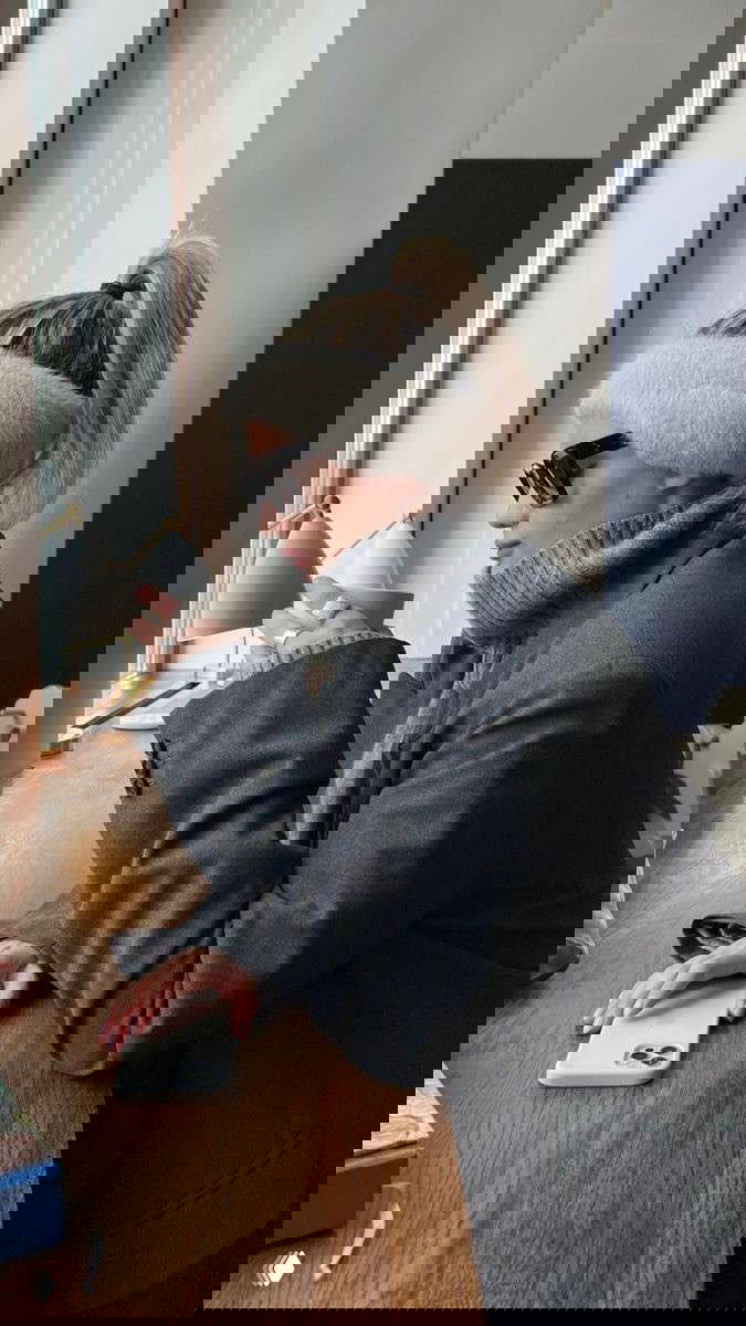 Профиль женщины в светлой пушистой повязке-наушнике и тёплом пальто, сидит у окна с чашкой на столе, образ в городском кафе.