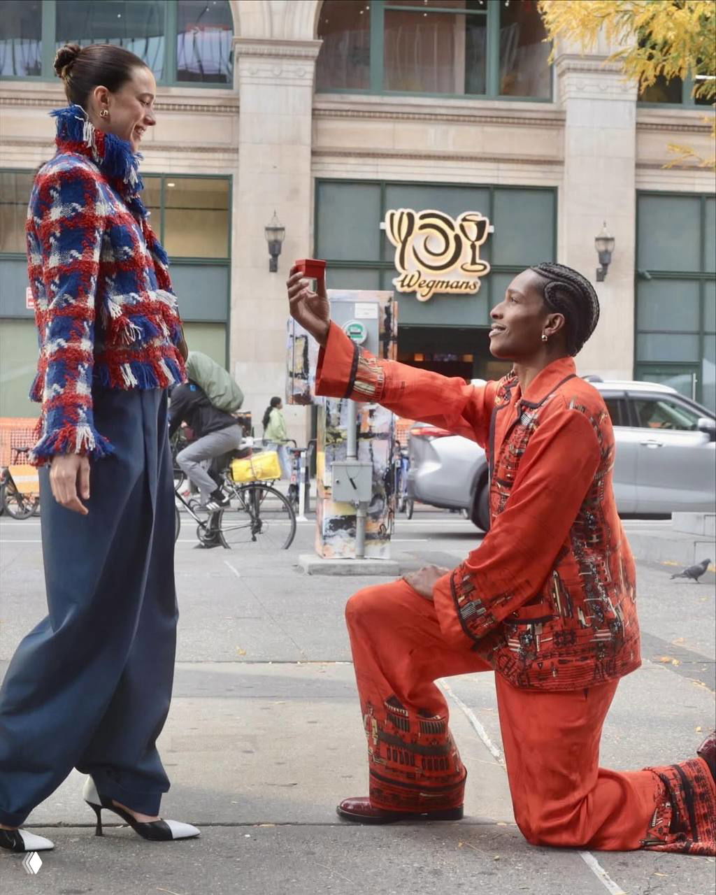 Мужчина на колене делает видимость предложения рядом с женщиной в стильной одежде на улице — кадр рекламной съёмки в городском окружении.