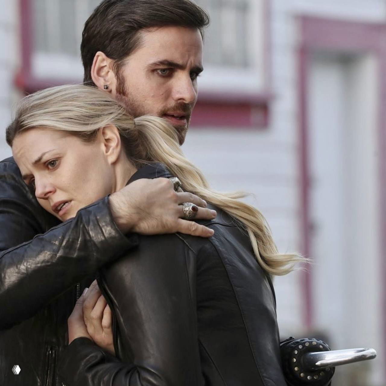 Крупный кадр: персонажи Эмма и Киллиан из сериала «Однажды в сказке» в крепком объятии на фоне городского фасада, напряжённые лица и кожа курток.