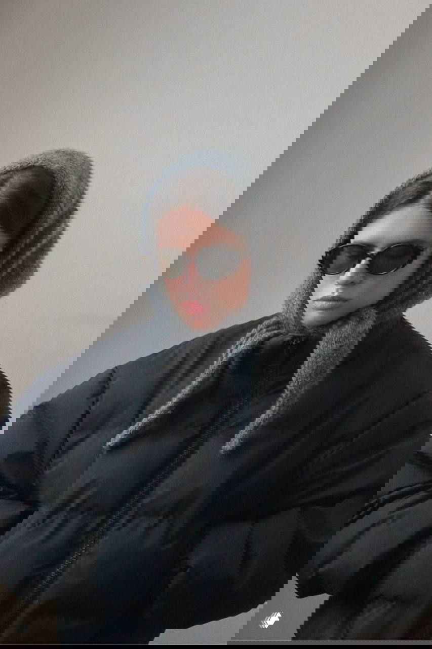 Модель в тёмной стёганой куртке и светлом платке-шапке, завязанном под подбородком; нейтральный студийный фон и солнцезащитные очки.