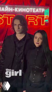 Два гостя в тёмной одежде позируют на фоне неоновой стены с надписями; кадр с красной подсветкой и логотипом СМИ на премьере