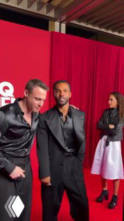 Крупный план двух мужчин в одинаковых черных шелковых рубашках на фоне красной стены с логотипом GQ; сценический кадр с гостями.