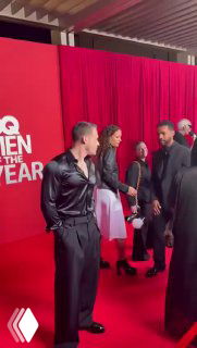 Два мужчины в черных шелковых рубашках на красной дорожке GQ Men of the Year; красная драпировка и логотип на фоне.