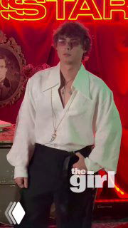 Мужчина в белой рубашке на фоне неоновой вывески и красной декорации с логотипом СМИ; кадр с премьеры, вечерняя сцена.