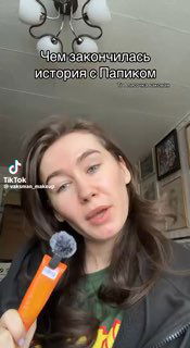 Кадр из видео: женщина держит в руках небольшой портативный микрофон/диктофон, на лице выражение рассказчика, фон — интерьер квартиры.