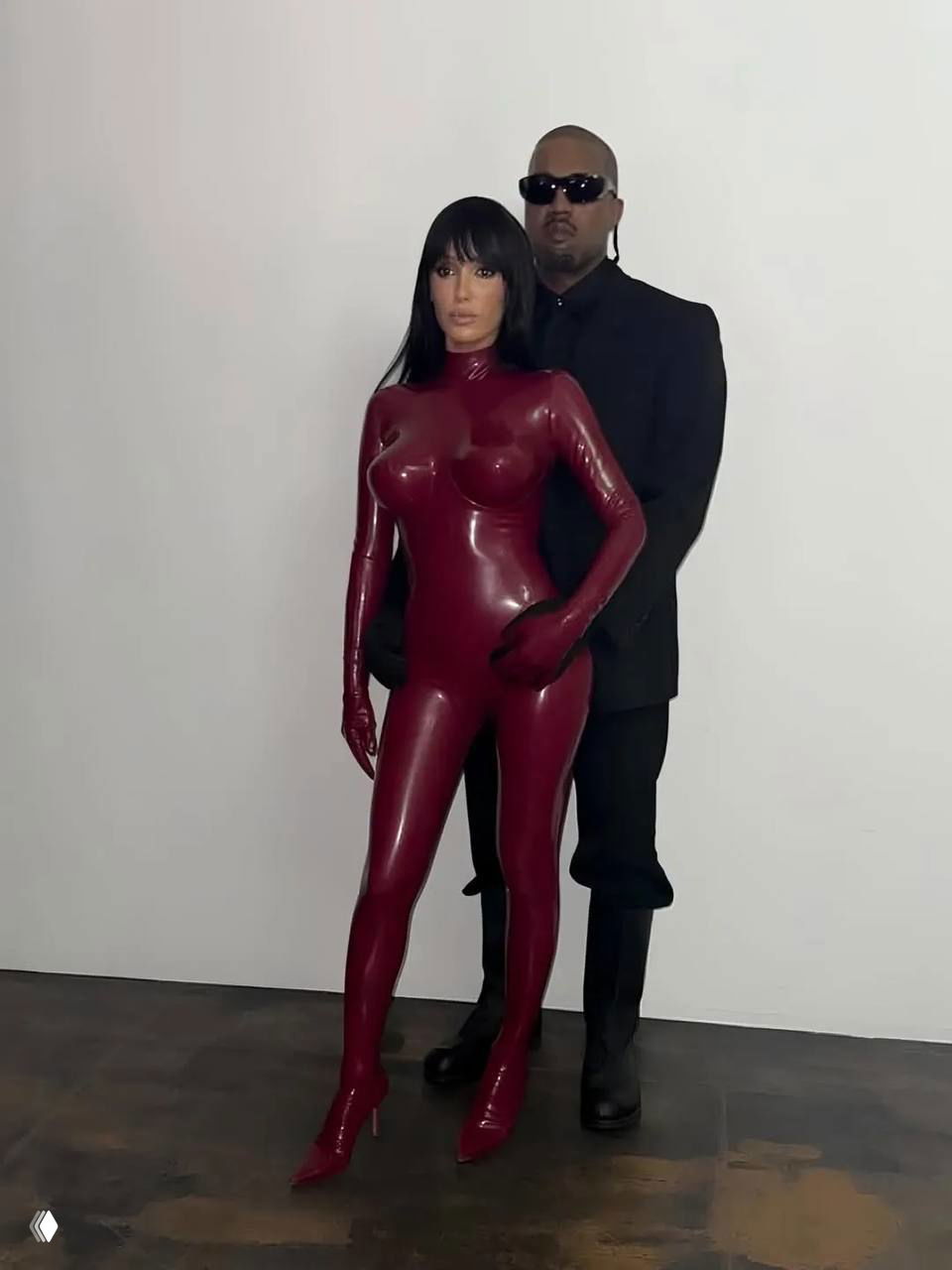 Бьянка Цензори в красном латексе на фоне белой стены, позади неё стоит Каньe Уэст в тёмной одежде — пара наблюдает за перформансом.