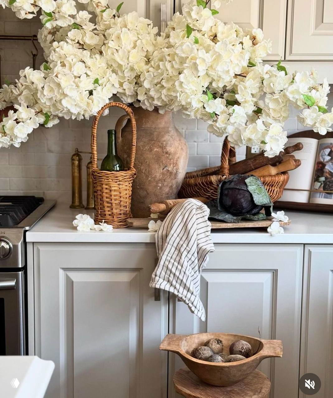 Кухонный натюрморт с большой вазой белых цветов, плетёной корзиной и деревянными аксессуарами, создающими уютную атмосферу интерьера.