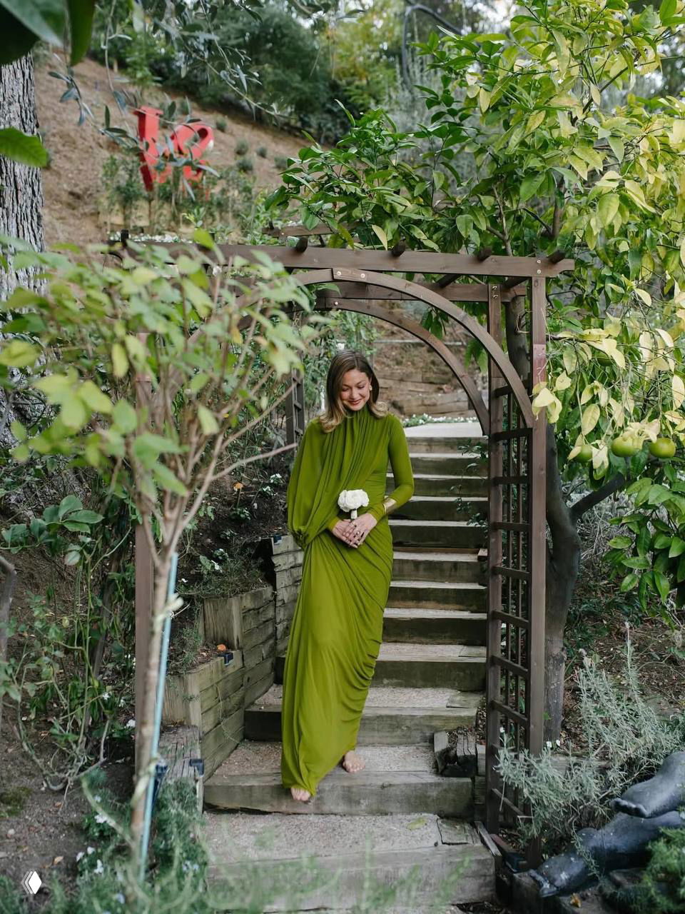 Женщина в длинном салатовом платье спускается по садовой лестнице под деревянной аркой, в руках у неё маленький белый букет, вокруг зелёная растительность.