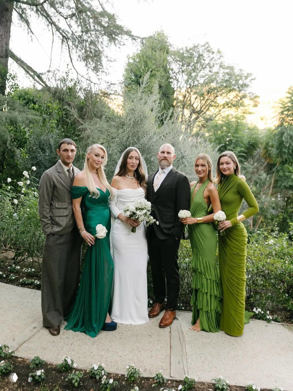 Групповая свадебная фотография в саду: невеста в белом платье в центре, рядом гости и подружки невесты в разных оттенках зелёного, мужской костюм.