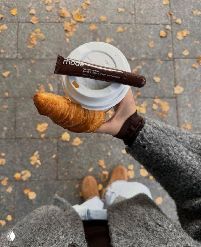 Снимок сверху: рука держит чашку кофе и круассан, на крышке лежит тюбик тинта для губ; осенние листья на тротуаре вокруг.