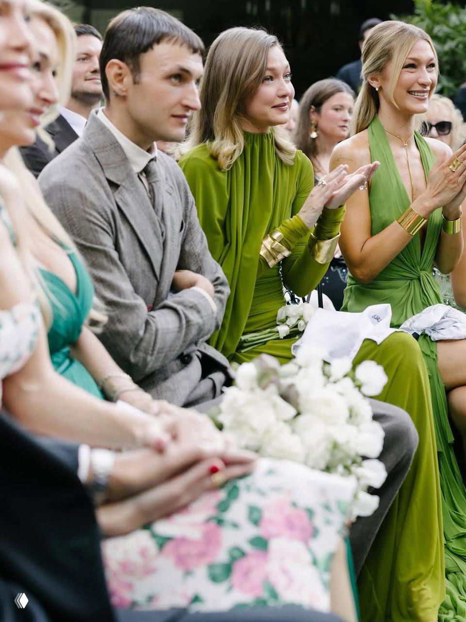 Крупный план свадебной аудитории: гости в рядах, две женщины в зелёных платьях аплодируют, в кадре видны белые букеты и элементы декора на переднем плане.