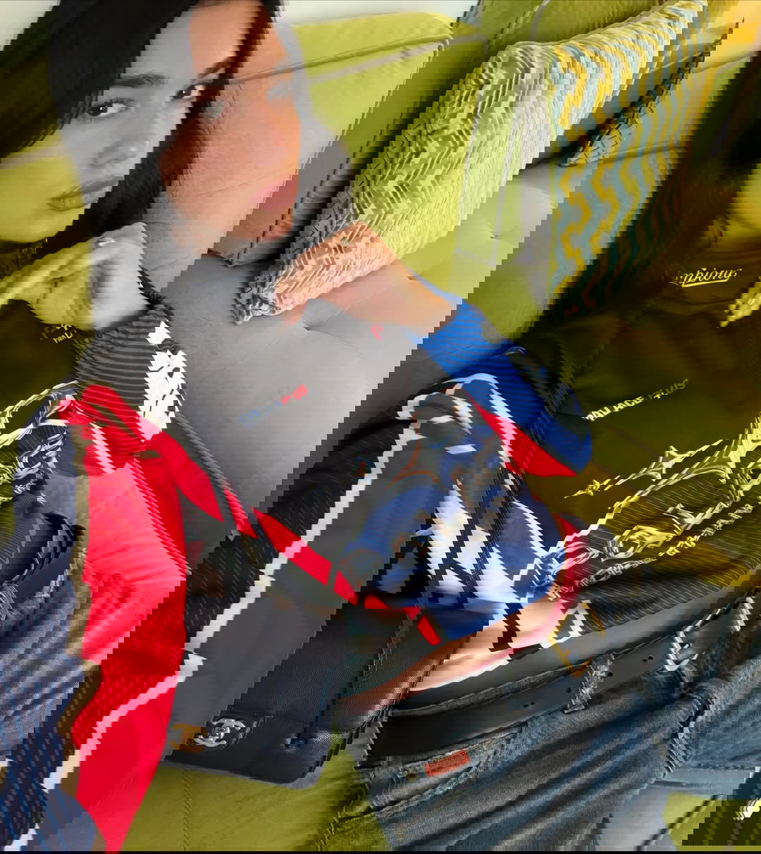 Портрет женщины в уличном образе: укороченный гоночный топ, джинсы и кожаная сумка, сидит на зелёном диване в помещении.