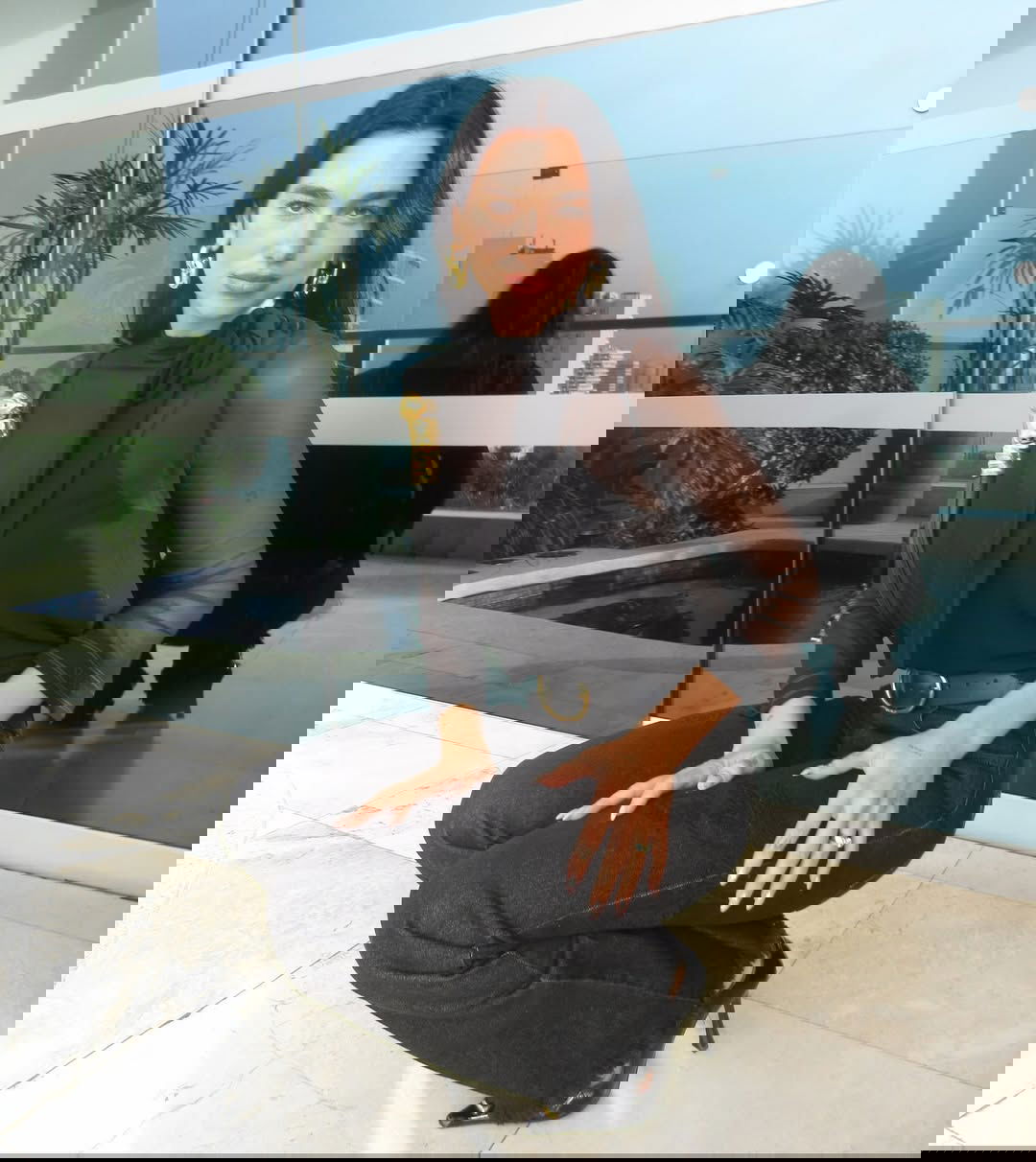 Женщина в элегантном чёрном образе: прозрачный топ и чёрные джинсы, позирует на балконе с частичным видом на город.