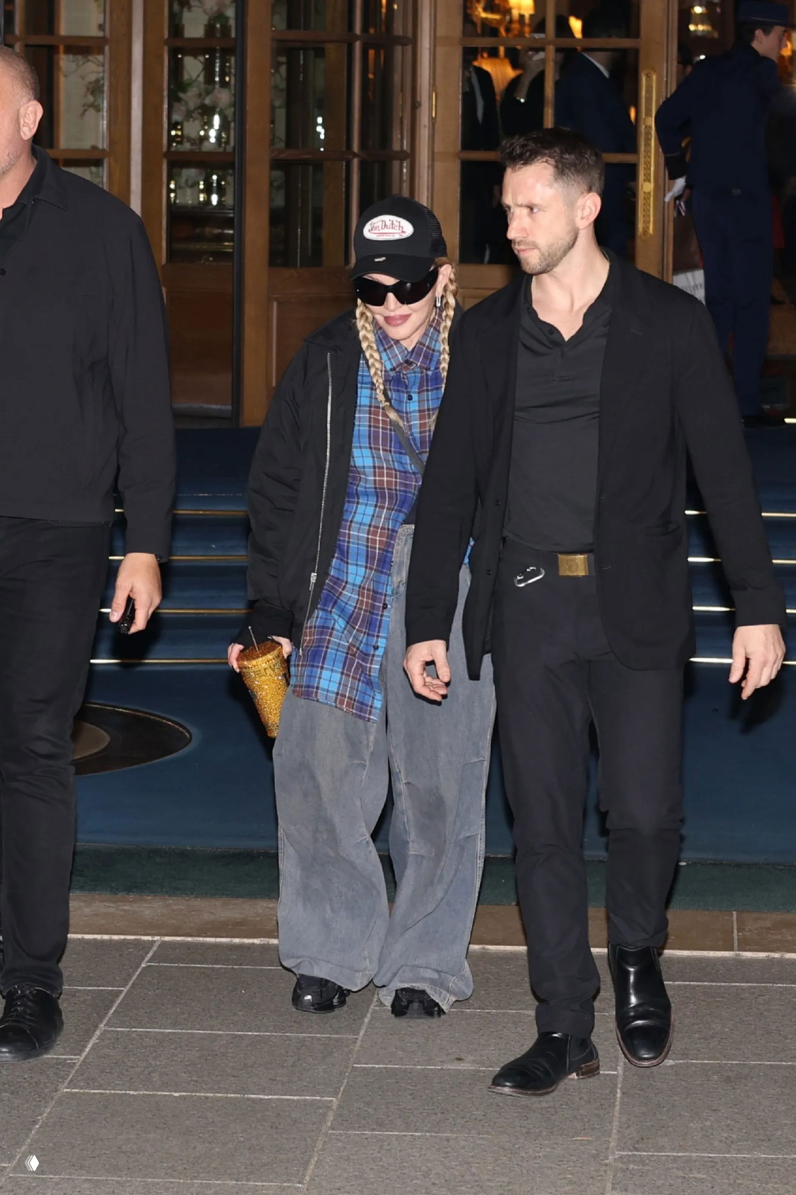 Женщина в тёмной кепке выходит из здания в сопровождении мужчины, уличный папарацци-снимок с акцентом на кепку и образ.