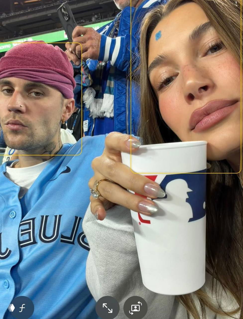 Селфи Джастина и Хейли на бейсбольном матче: Хейли ближе к камере с чашкой в руках, Джастин в розовой шапке и синей футболке на заднем плане.