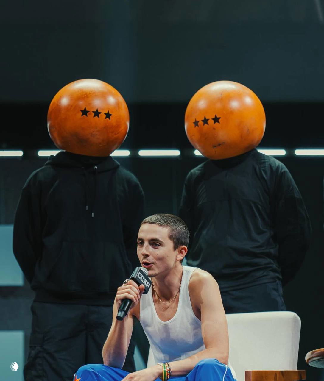 Мужчина с короткой стрижкой сидит на сцене и держит микрофон; за ним два человека в чёрной одежде с круглыми масками‑шлемами — сценический кадр интервью или презентации.