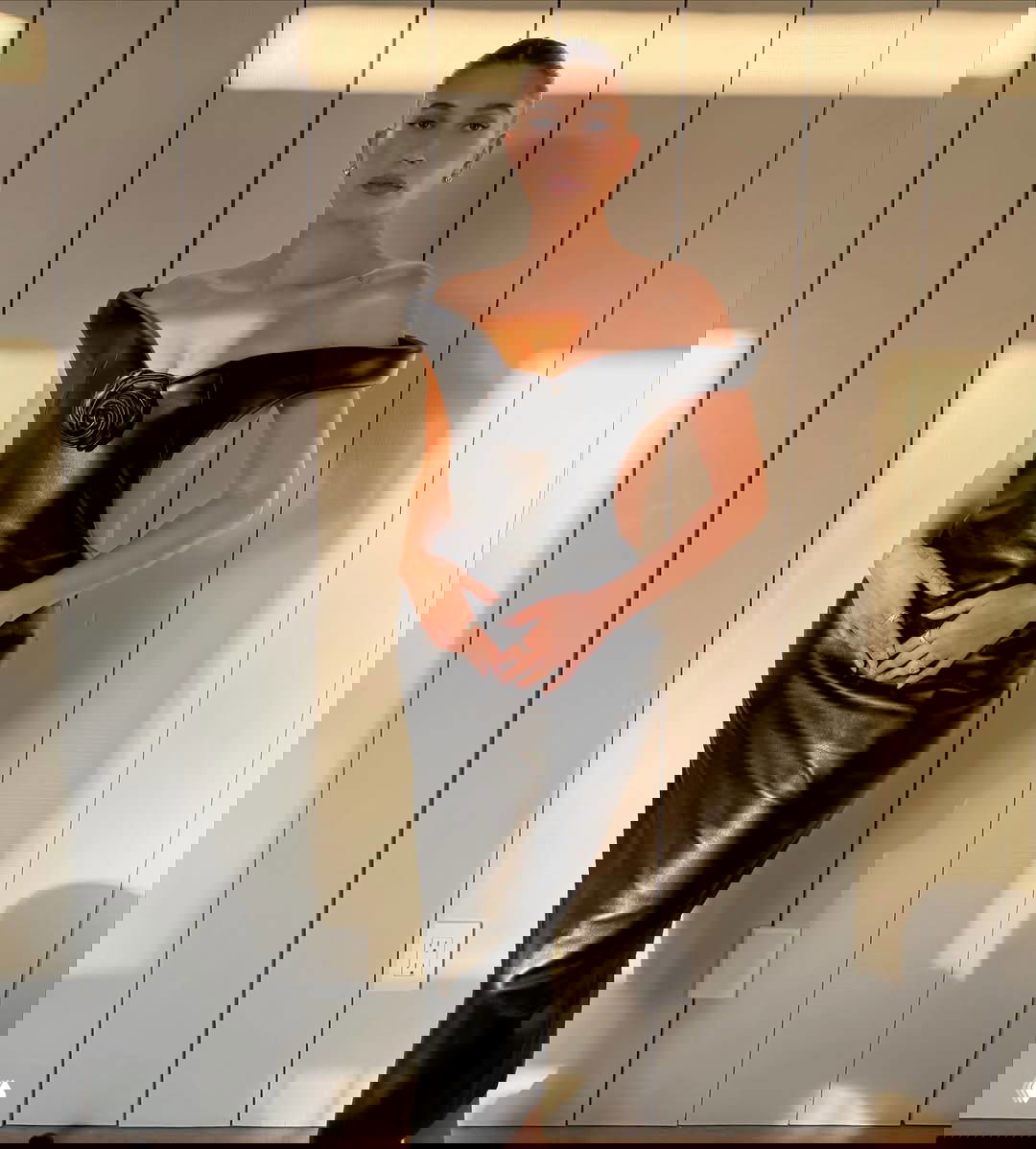 Женщина в облегающем чёрном кожаном платье с открытыми плечами и крупным цветочным декором у груди, позирует на нейтральном фоне.