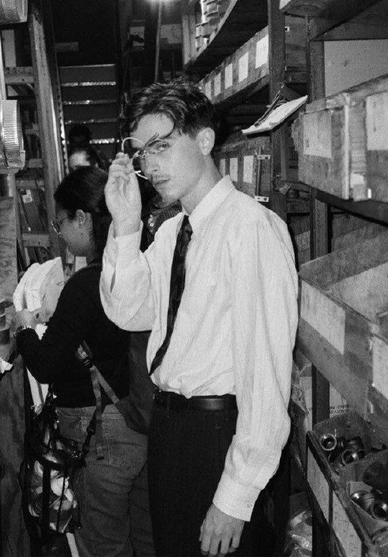 Чёрно‑белое фото мужчины в очках и рубашке в складском помещении; атмосферный кадр среди стеллажей и упаковок, задумчивый портретный ракурс.