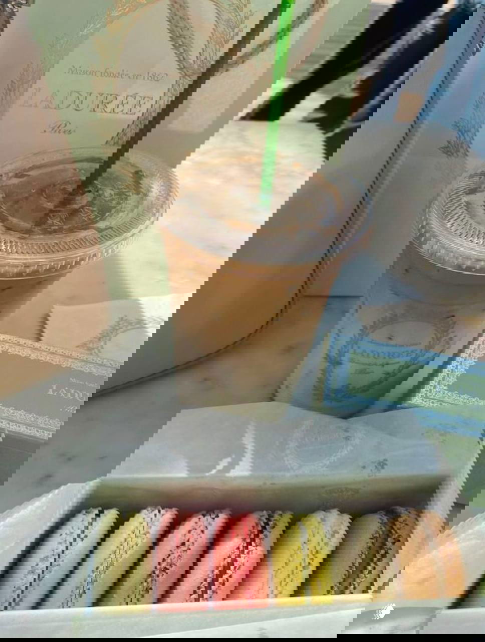 Коробка макарунов и стакан охлаждённого кофе на мраморном столе — пастельный натюрморт с фирменной упаковкой кондитерской.