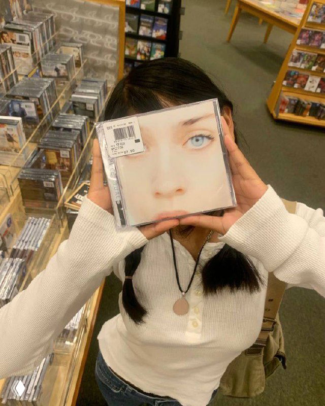 Девушка с косичками держит CD‑коробку с большим портретом перед лицом в магазине, на заднем плане видны полки с дисками.