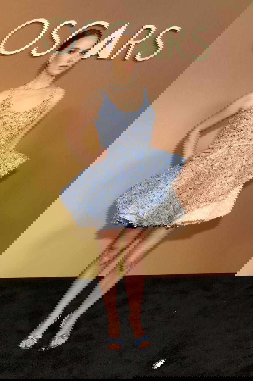 Женщина в коротком голубом фактурном платье на красной дорожке Governors Awards, позирует в полный рост на фоне логотипа церемонии, фотография с Governors Awards в Лос-Анджелесе.