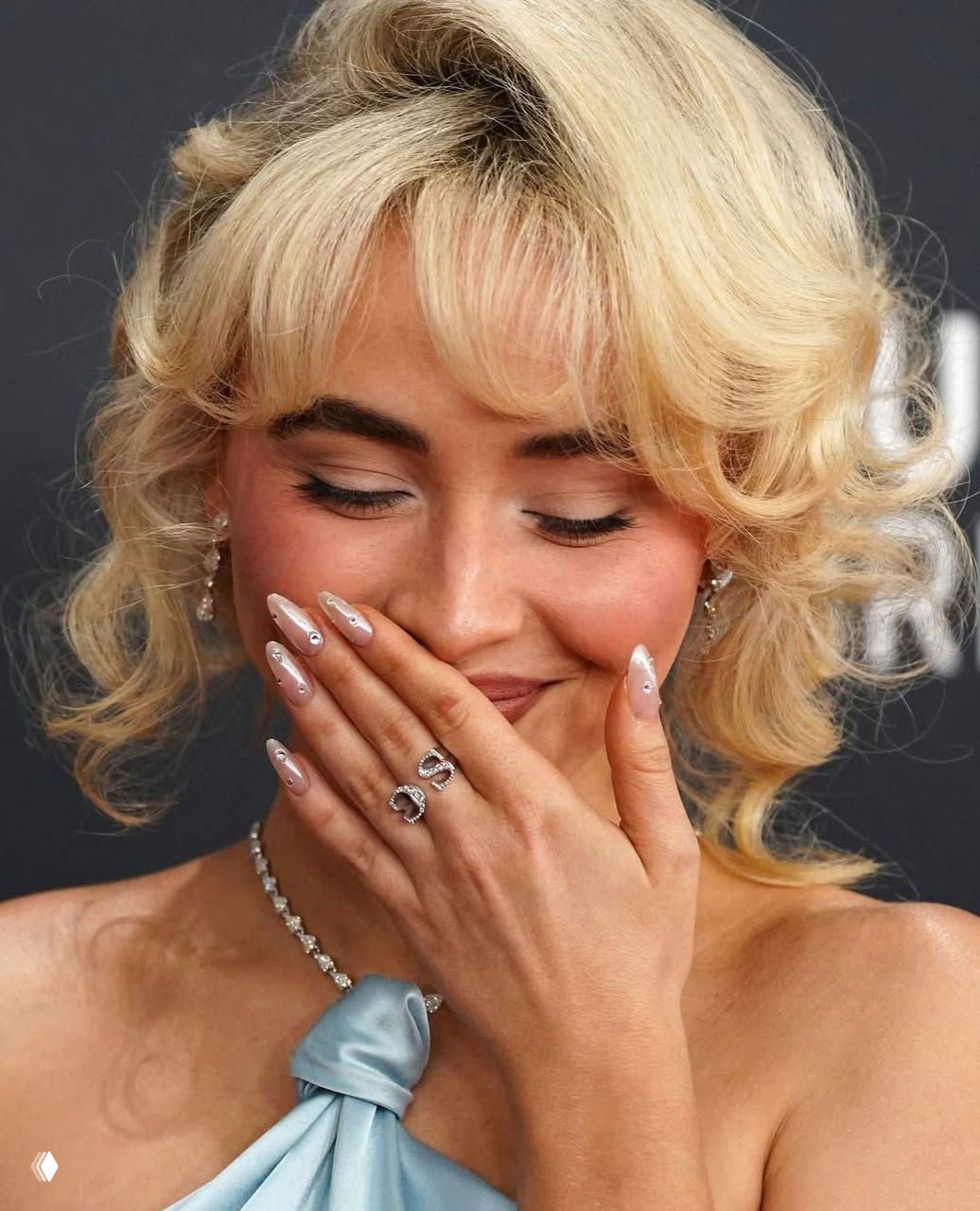 Портрет: женщина прикрывает рот рукой; видны длинные нюдовые ногти с жемчужными акцентами и изящные кольца на пальцах.