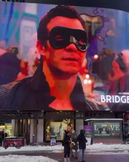 Биллборд с кадром из «Бриджертонов» над заснеженным тротуаром и прохожими — реклама сериала на городской улице рядом с витринами магазинов.