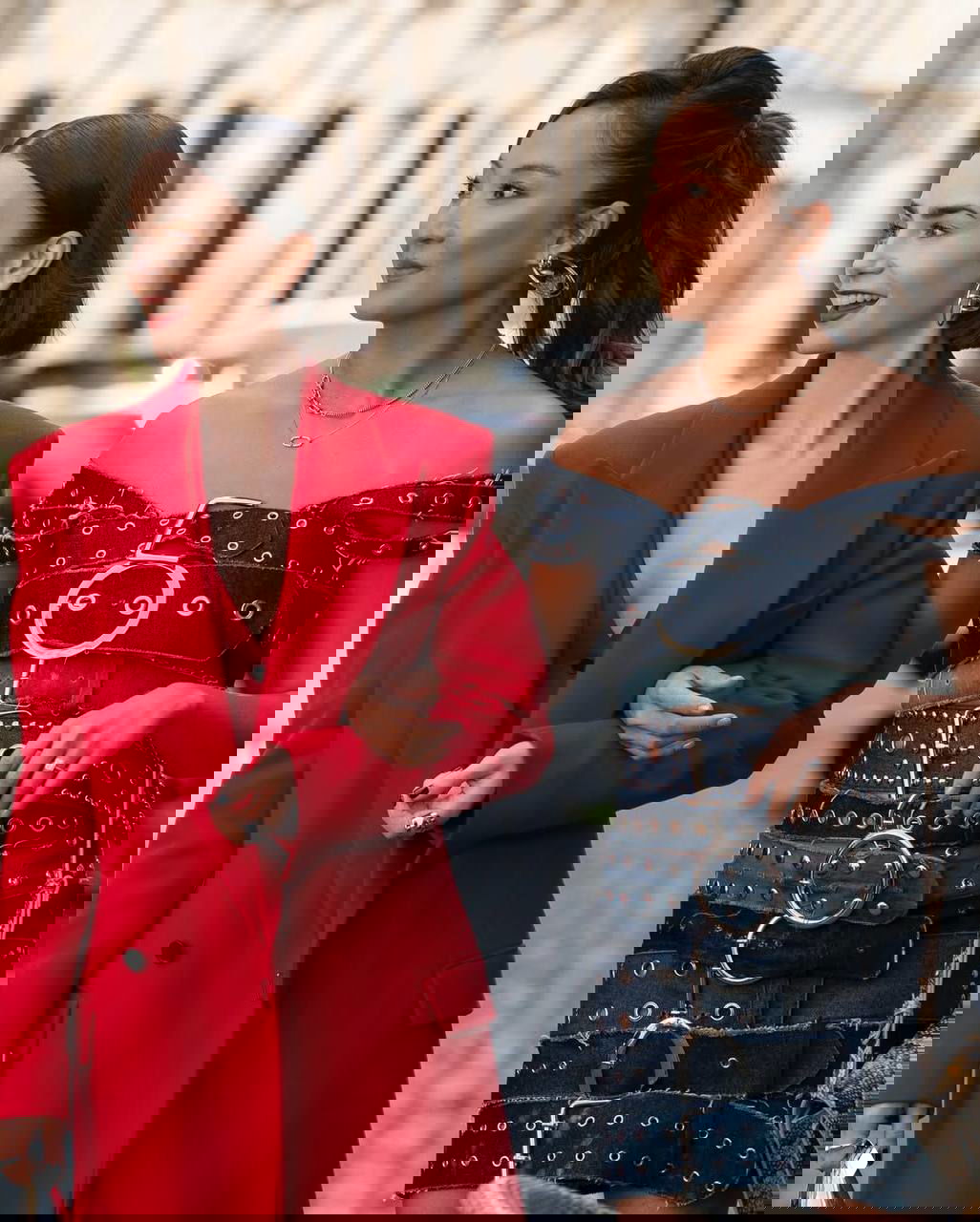 Две женщины в стильных образах идут по улице: одна в ярком красном пальто, вторая в джинсовом платье с декоративными ремнями; акцент на моде и аксессуарах.