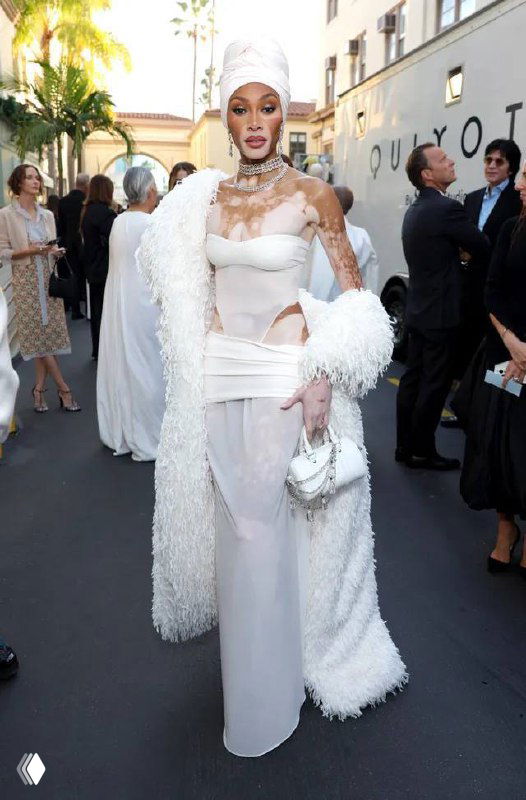 Знаменитость в белом облегающем платье со структурным элементом и пушистым манто позирует на красной дорожке у входа в мероприятие.