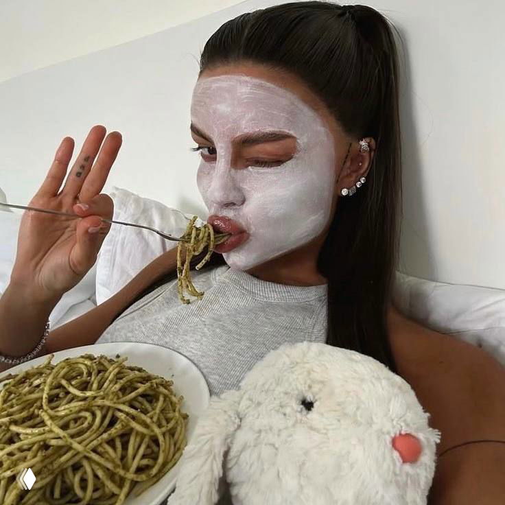 Девушка в маске для лица ест пасту на диване — игривый кадр в стиле self-care и домашнего отдыха во время праздников.