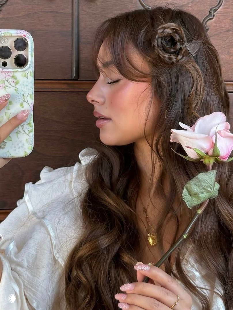 Портрет: девушка с волнистыми волосами держит розу и телефон с цветочным чехлом; видны аккуратный маникюр и золотая подвеска.