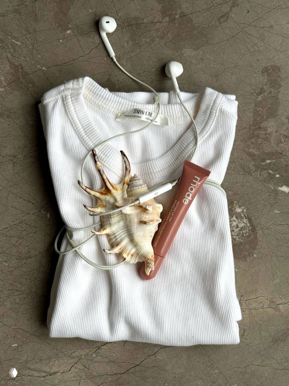 Сложенный белый топ с наушниками, ракушкой и тюбиком помады — аккуратная бьюти-композиция в нейтральных пастельных тонах