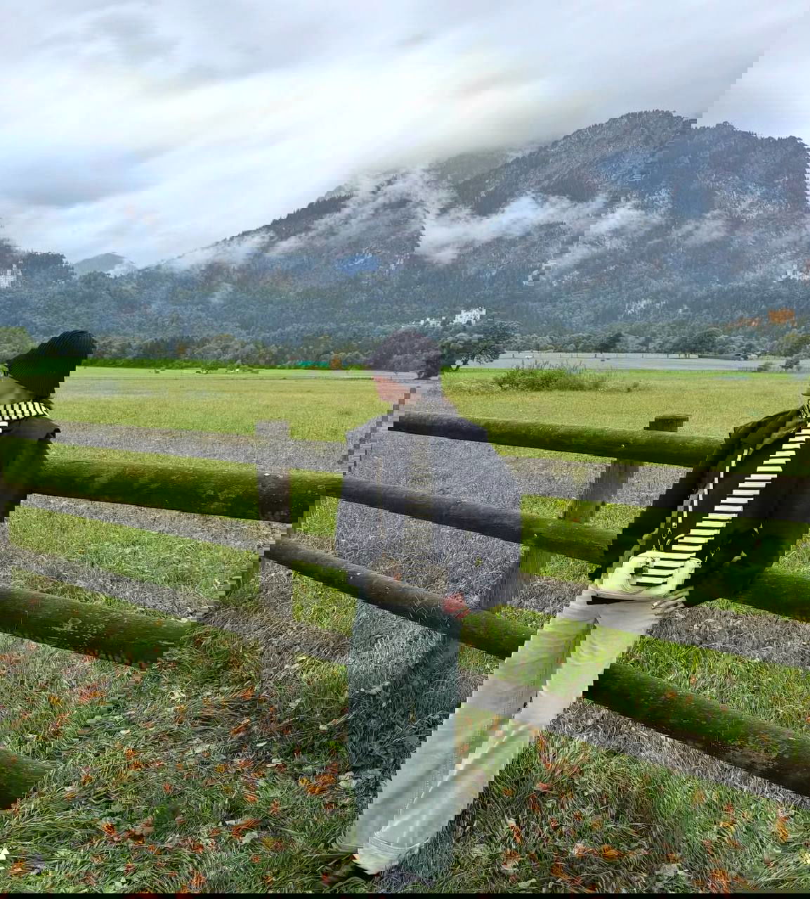 Лиза у деревянного забора на фоне горного ландшафта: черная шляпа клош, полосатый шарф и свободные светлые брюки в осеннем антураже.