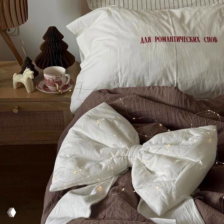 Крупный план кровати с бантом-подушкой и постельным бельём, украшенным мелкими огоньками — романтичный праздничный декор спальни.