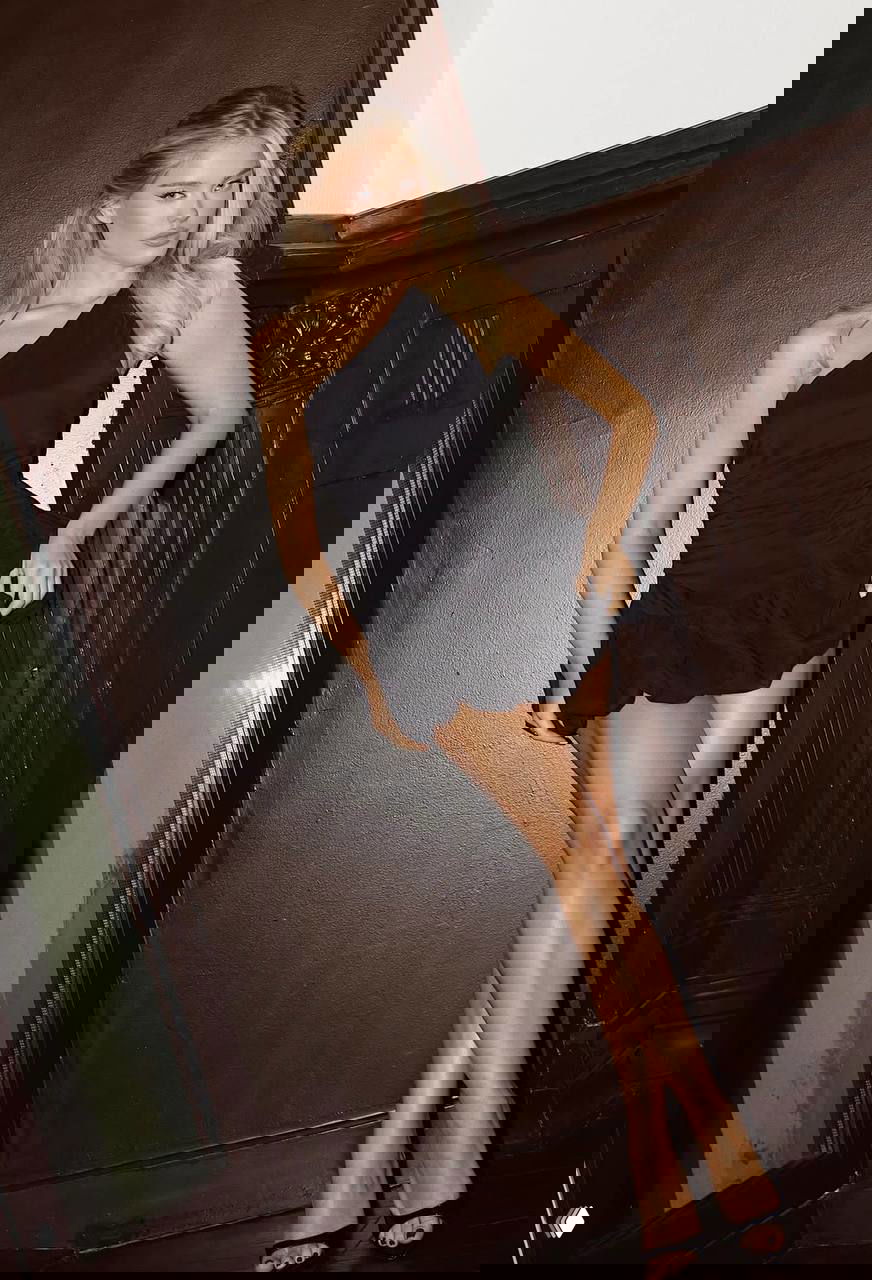 Катя Мотовилова в чёрном асимметричном платье у стены — элегантный образ с открытыми ногами и чёрными каблуками, студийная или интерьерная съёмка.