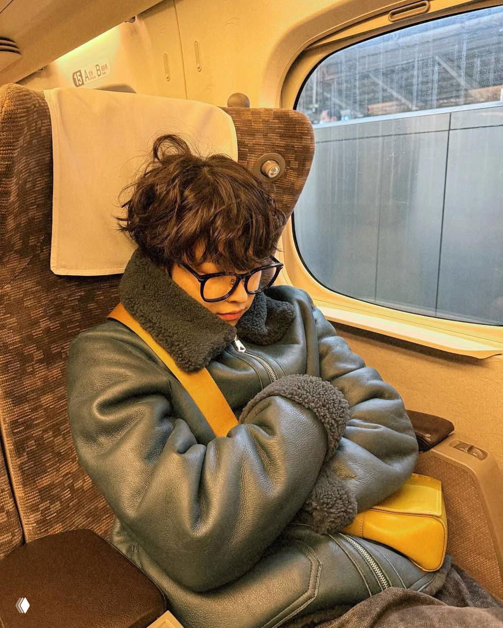 Хонджун в тёплой дублёнке и очках, сидит и дремлет в кресле поезда рядом с окном, видно сумку через плечо и интерьер вагона.