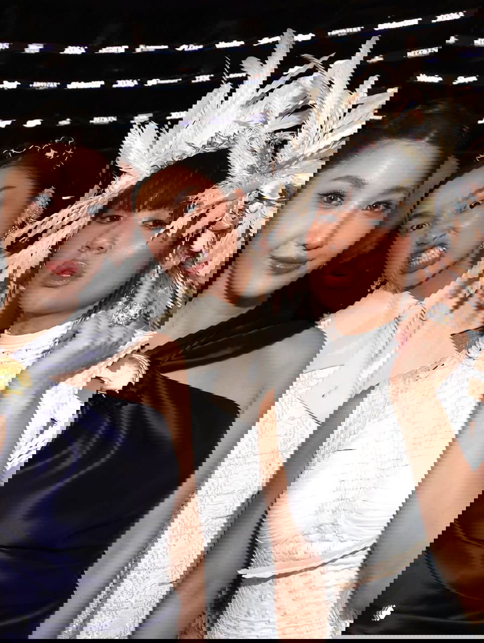 Группа гостей в стильных нарядах позирует рядом друг с другом; заметны сложные головные украшения и вечерние платья.