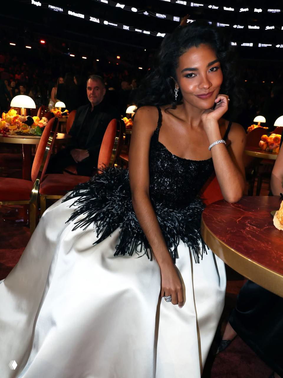 Женщина в чёрно‑белом вечернем платье сидит за столом в зале церемонии, улыбается и опирается на руку; вокруг лампы и декор.