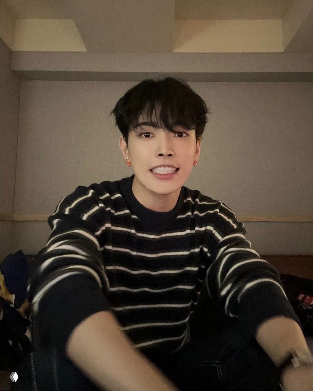 Селфи Хонджуна в тёмной комнате: тёмные волосы, полосатый свитер и улыбка в камеру, уютный студийный фон за спиной.