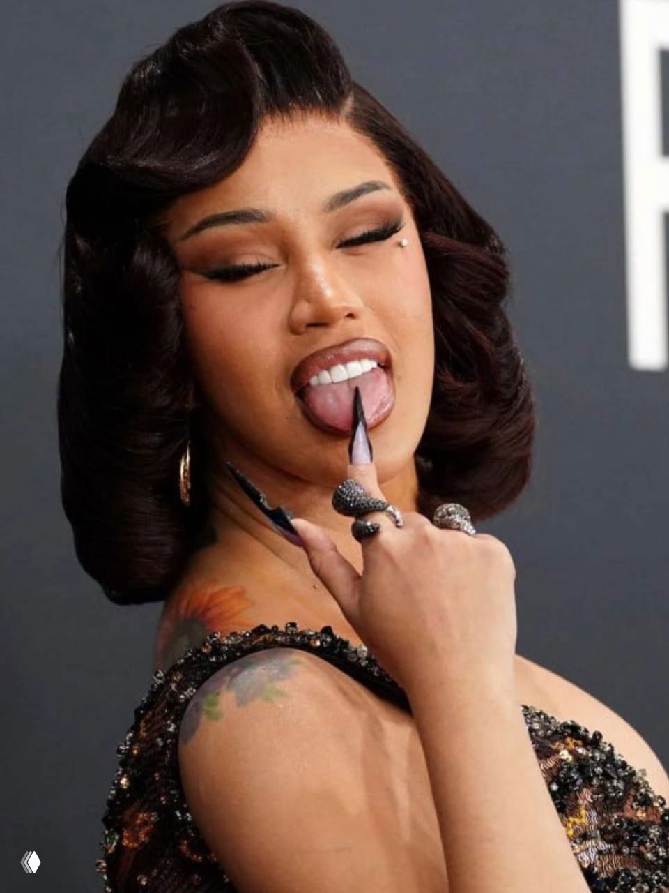 Артистка в крупном плане показывает язык и длинные ногти, видна татуировка на плече, игривый жест и выразительный макияж.