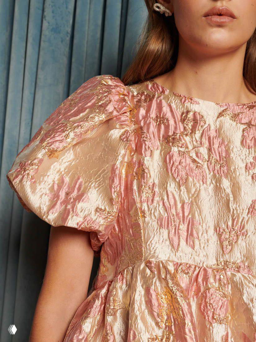 Крупный план пастельного жаккарда: платье с пышным рукавом, золотистые и розовые переливы, тиснёный узор и текстура ткани на фоне занавесок.