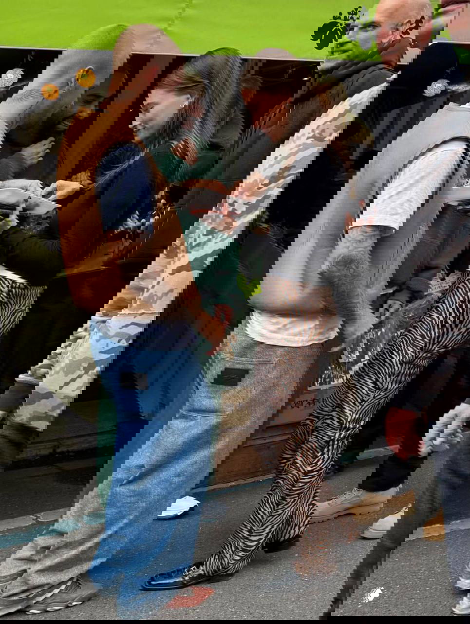 Папарацци‑кадр у торговой палатки: пара рассматривает коробку с уличными закусками, женщина в зебровых брюках пробует еду, вокруг — рынок и люди.